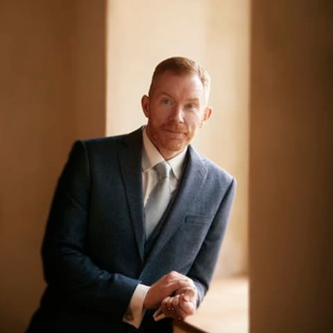 James Morrison Concert One | Brass Sonority - Man in a blue-grey blazer stands in warm light from a window, hands clasped thoughtfully before him.
