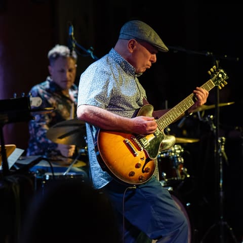 Miles Davis at 100 | Curated by Carl Orr - Guitarist in blue cap and shirt playing sunburst electric guitar on stage, with ensemble members visible behind in dramatic stage lighting.