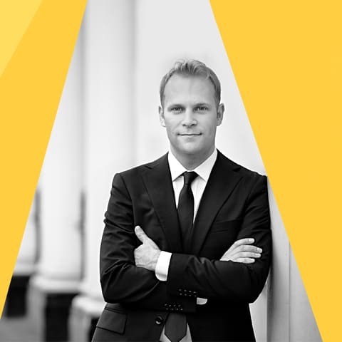 Chamberfest Concert One | Origins - Man in dark suit with arms crossed against geometric yellow and white backdrop.