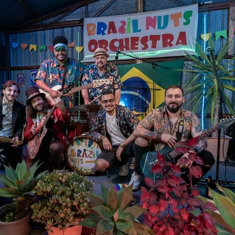 Brazil Nuts Orchestra - Brazil Nuts Orchestra members pose together in front of their illuminated band name sign at Payinthi venue, surrounded by potted plants.