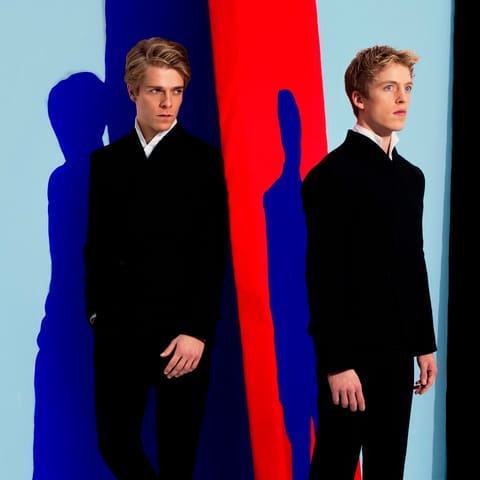 Lucas and Arthur Jussen - Two pianists in dark suits stand against a colourful abstract backdrop of blue, red and yellow panels.