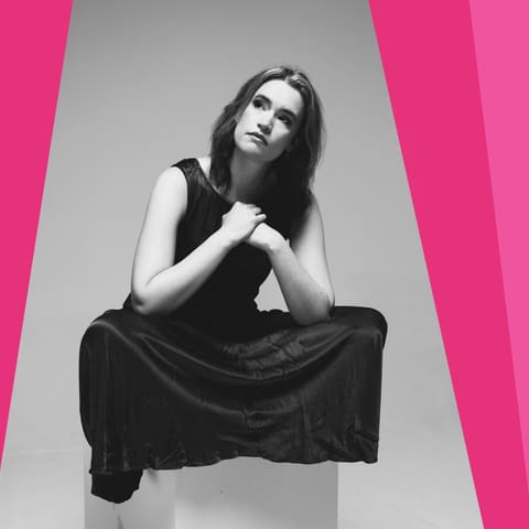 Chamberfest Concert Four | The Mighty and Mundane - Woman in black clothing seated against pink and grey geometric backdrop, looking upward thoughtfully.