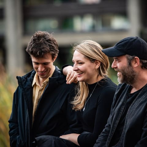Za Górami - Three performers stand together outdoors at UKARIA Cultural Centre, smiling in casual dark clothing against a blurred industrial backdrop.