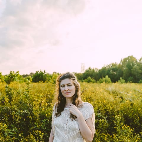 Ruth Moody | The Wanderer Returns - Woman in a white dress standing in a golden wildflower meadow under cloudy sky.