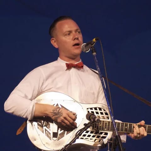 Musician performing with white resonator guitar and red bow tie against blue stage backdrop.