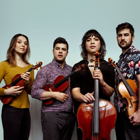 Attacca Quartet - Four musicians from Attacca Quartet stand together holding their instruments against a pale blue background.