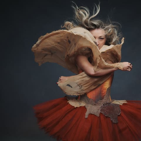 Kate Miller-Heidke - Performer in a red tutu and brown fabric mid-leap against a dark background.