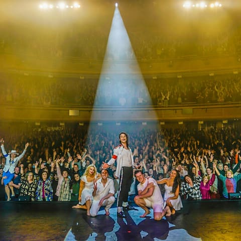 The King of Pop Show - A performer stands spotlit centre stage whilst a packed audience fills Her Majesty's Theatre around them.