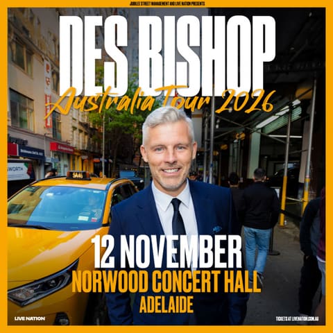 Des Bishop - Australia Tour - Man in dark suit and tie smiling at camera on colourful city street with yellow taxi visible.