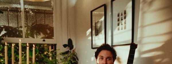 Plini ‘An Unnameable Desire’ Australia & New Zealand Tour - Musician sits at a table with a small dog in a warmly lit interior space with plants and framed artwork.