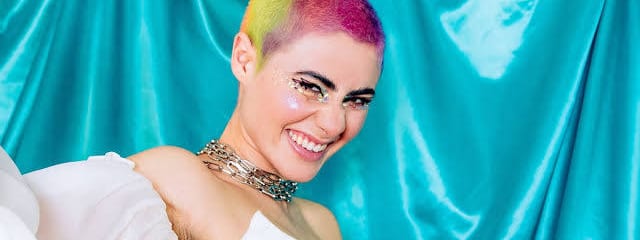 Montaigne - Glorious Heights 10th Anniversary Tour - Person with vibrant rainbow hair and gold chain jewellery against a turquoise fabric backdrop.