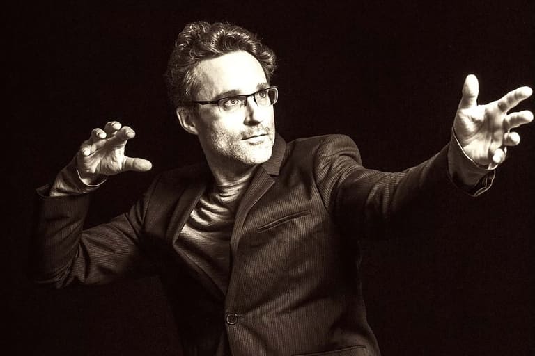 Man in dark suit and glasses gestures expressively against black background during performance.
