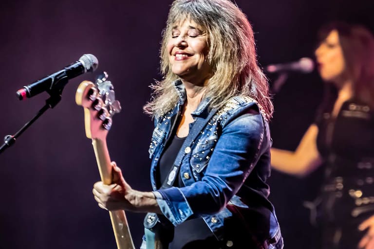 SUZI QUATRO - Musician in a blue jacket plays bass guitar at a microphone on stage with a band member visible in the background.