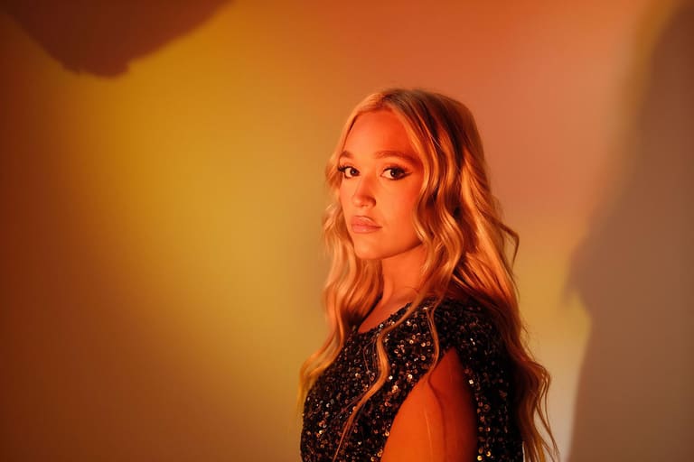 IVANA remakes Jennifer Paige and Cascada for the dancefloor - Performer with long blonde hair in a sequined black jacket against warm orange and yellow stage lighting.