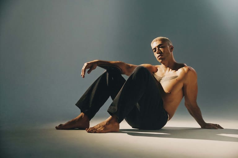 Teeks - Shirtless man seated on floor against grey background, wearing dark trousers and glasses.