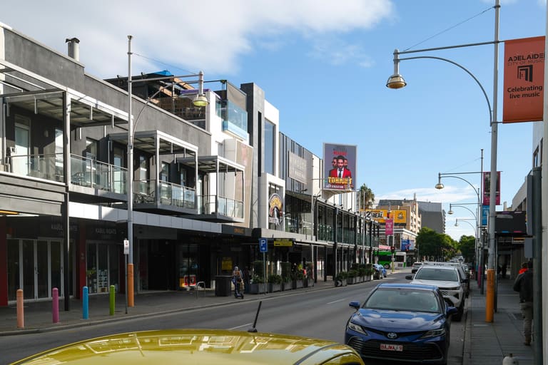 Hindley Street Music Hall - A street view of a commercial district with multi-storey buildings, shops, and parked cars under blue sky.