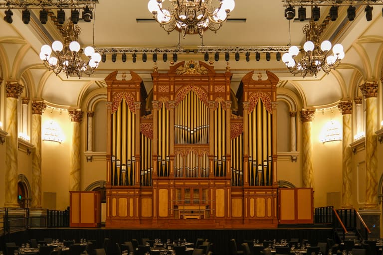 Adelaide Town Hall - A grand pipe organ with golden pipes dominates the ornate cream and gold interior of a classical concert hall.