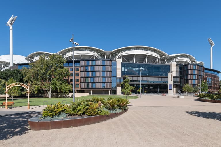 Adelaide Oval - Modern curved building with blue glass facade, landscaped courtyard, and tall white flagpoles under clear sky.