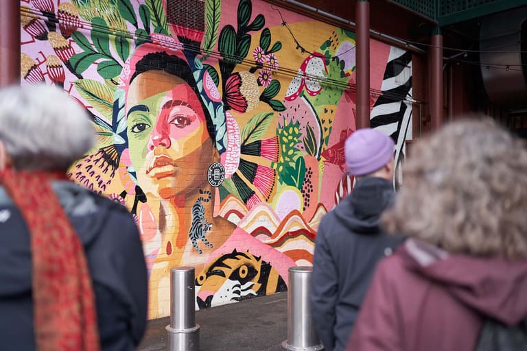 ,000 artists take over South Australia in August's SALA Festival - Gallery visitors view a vibrant portrait mural featuring a face painted in pink, green and yellow tones surrounded by botanical imagery.