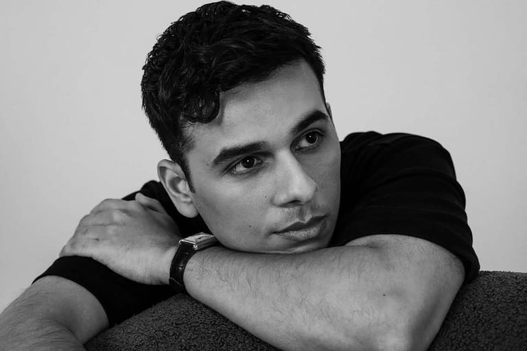 Man with dark hair resting his chin on his arms against a neutral grey background in black and white portrait.