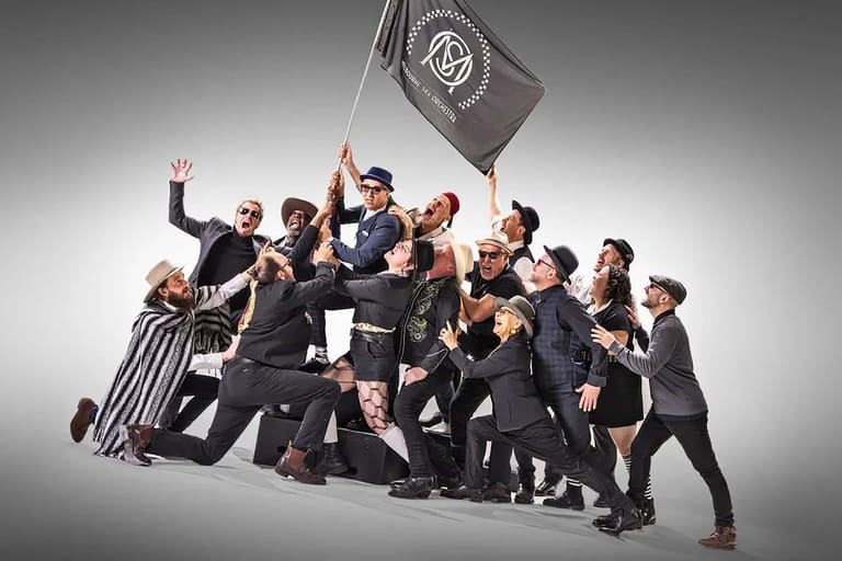 Group of performers in dark suits clustered together, one holding aloft a grey flag, against a neutral background.
