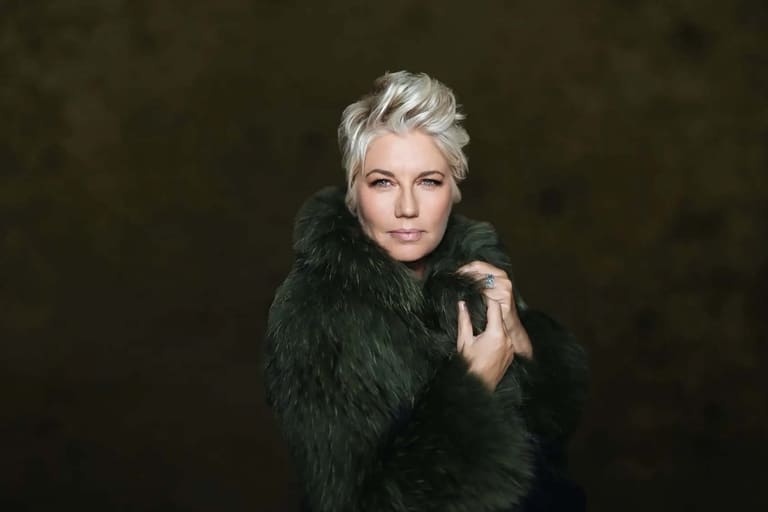 Person with platinum blonde hair wearing a black fur-collared coat against a dark background.