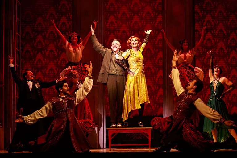 Two performers in vintage clothing stand centre stage with arms raised, surrounded by ensemble cast on a richly patterned red set.
