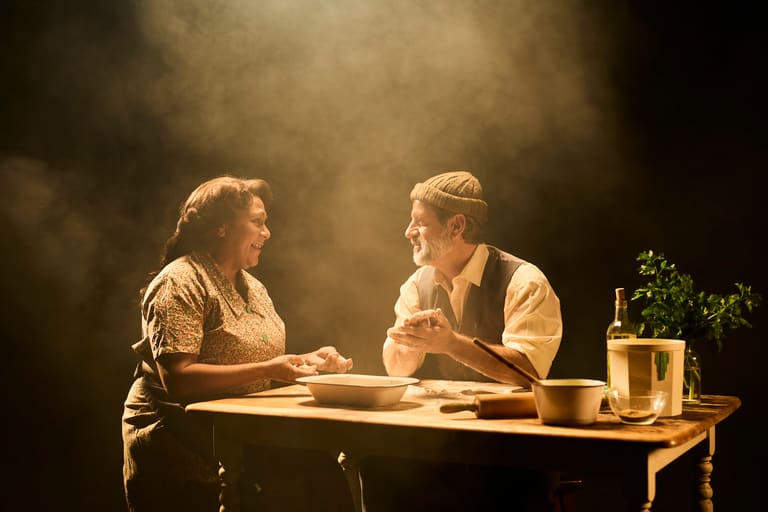 Two figures sit across a wooden table in warm stage lighting, engaged in intimate conversation.