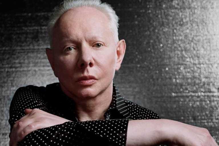 Musician with white hair in black polka-dot shirt against textured grey background, arms crossed.