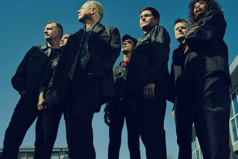 Five band members in dark suits stand against a blue sky, looking upward in a promotional pose.