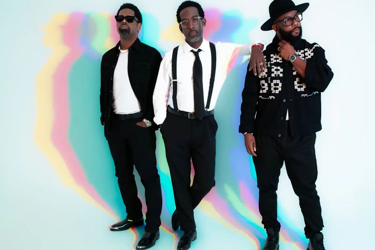Three men in black suits and sunglasses stand against a pastel rainbow gradient backdrop.
