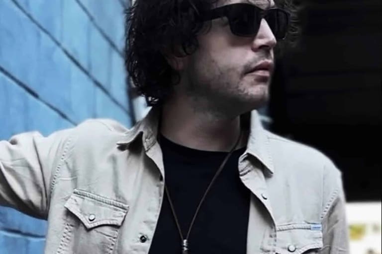 Man in sunglasses and light denim shirt against a blue building exterior.
