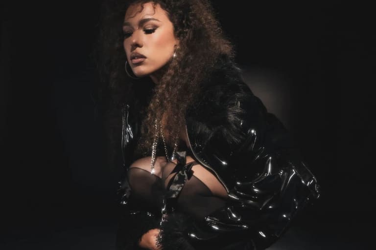 Person with curly dark hair in black leather jacket against dark background, gazing downward.