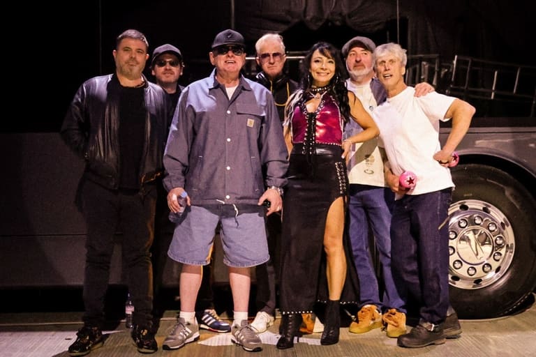 Seven band members stand together in front of a tour bus, dressed in casual 1990s-style clothing.