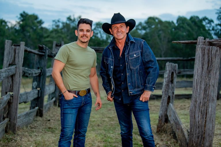 Two men in casual western wear standing by a weathered wooden fence in a rural paddock setting.