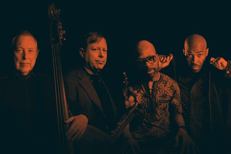 Five male musicians pose together holding instruments against a dark background.
