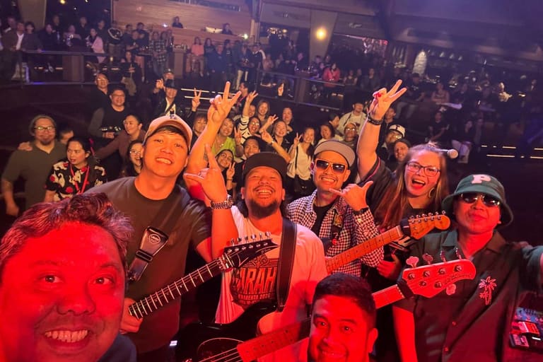 Crowd of enthusiasts in 90s attire dancing and cheering with arms raised at a concert venue under red stage lighting.