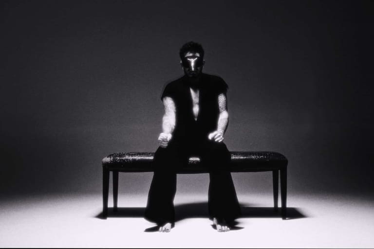 Person in black and white outfit sitting on a bench against a stark grey backdrop.