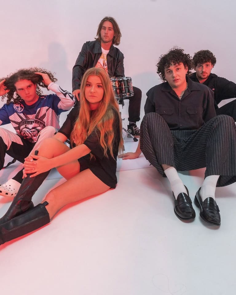 Five band members posed together against a white background lit by pink and red stage lighting.