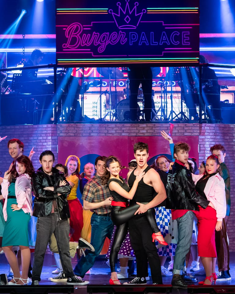 Large ensemble cast in colourful 1950s costumes performing on a brightly lit stage during Grease production.