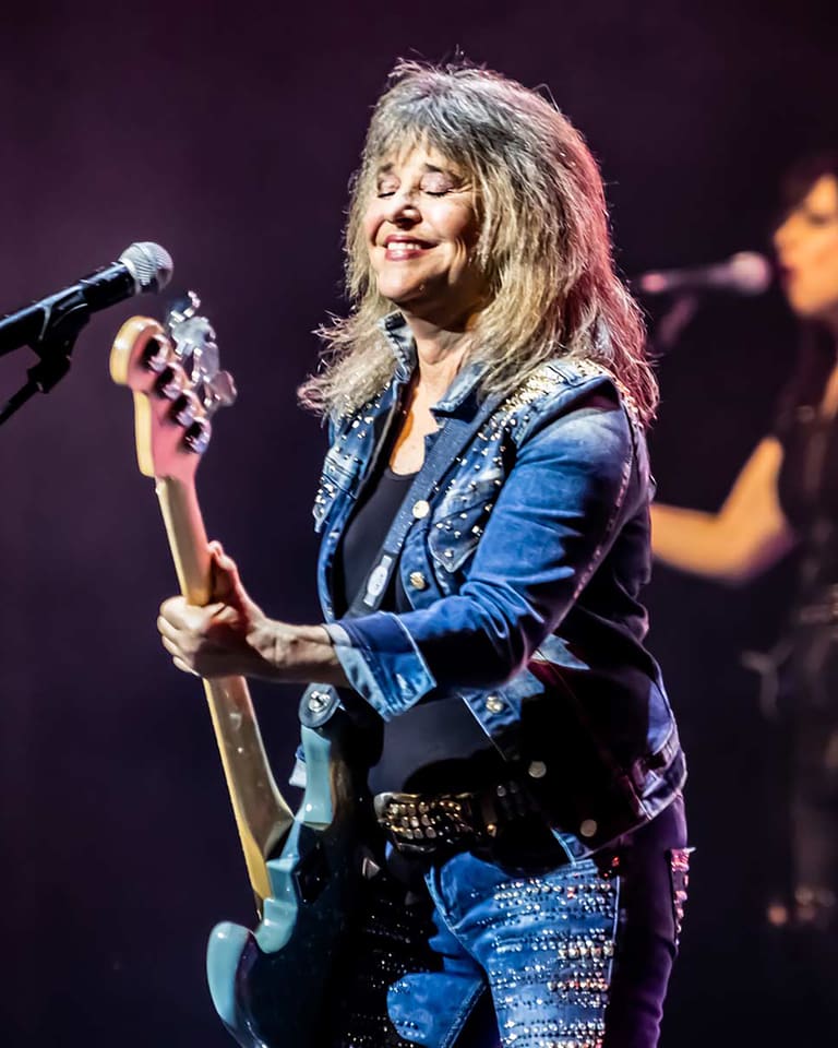 SUZI QUATRO - Musician in a blue jacket plays bass guitar at a microphone on stage with a band member visible in the background.