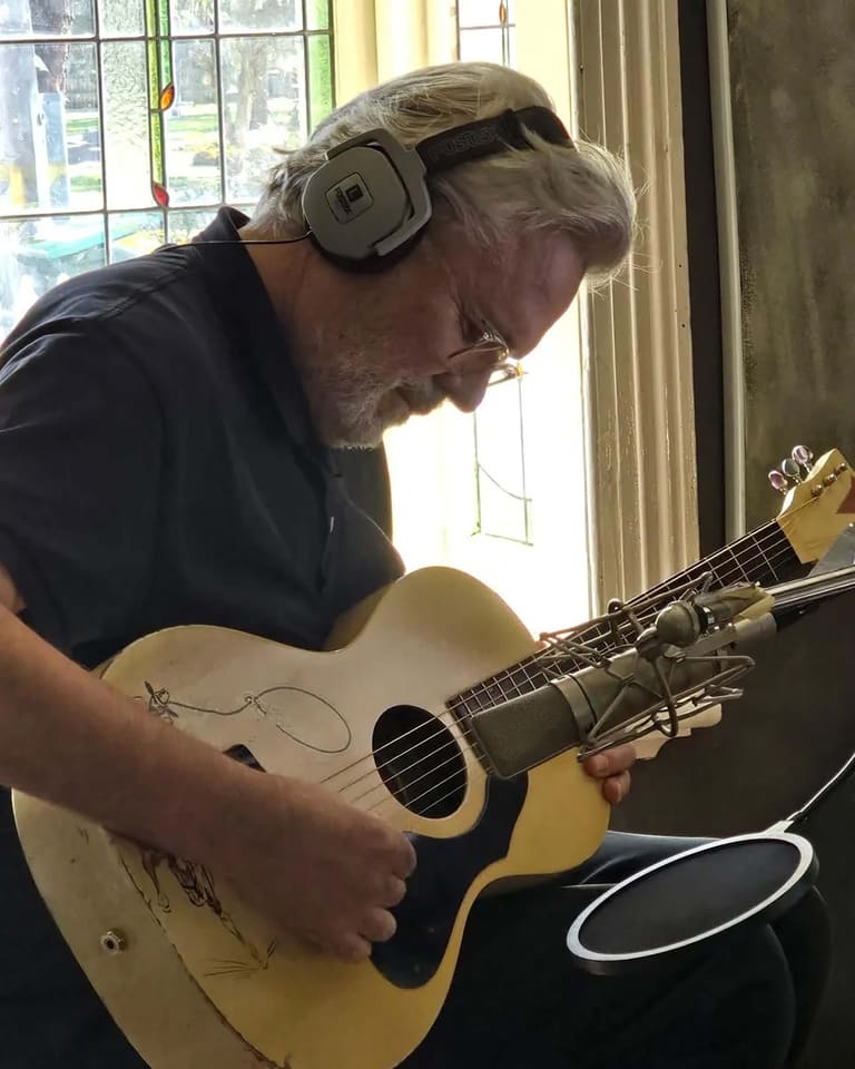 Musician with headphones around neck playing acoustic guitar indoors near a window.