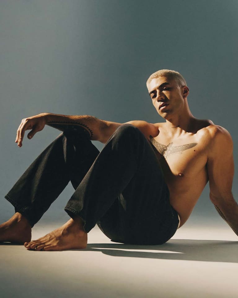 Teeks - Shirtless man seated on floor against grey background, wearing dark trousers and glasses.