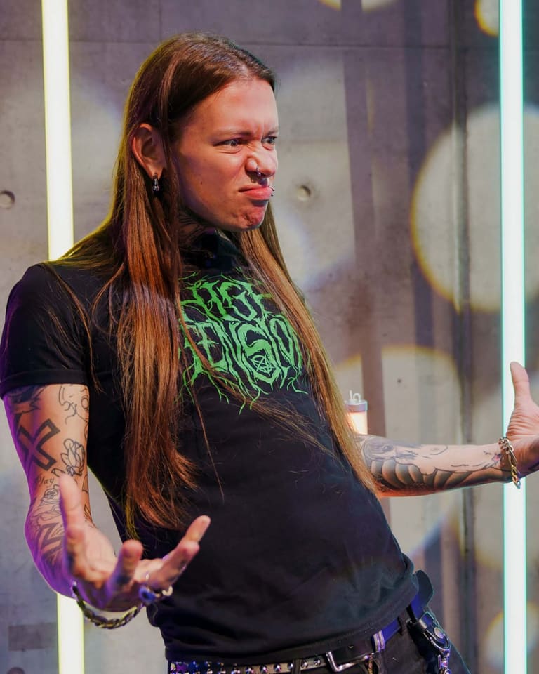 Nat’s What I Reckon - Performer with long brown hair and tattooed arms gestures expressively on stage during Nat's What I Reckon show.