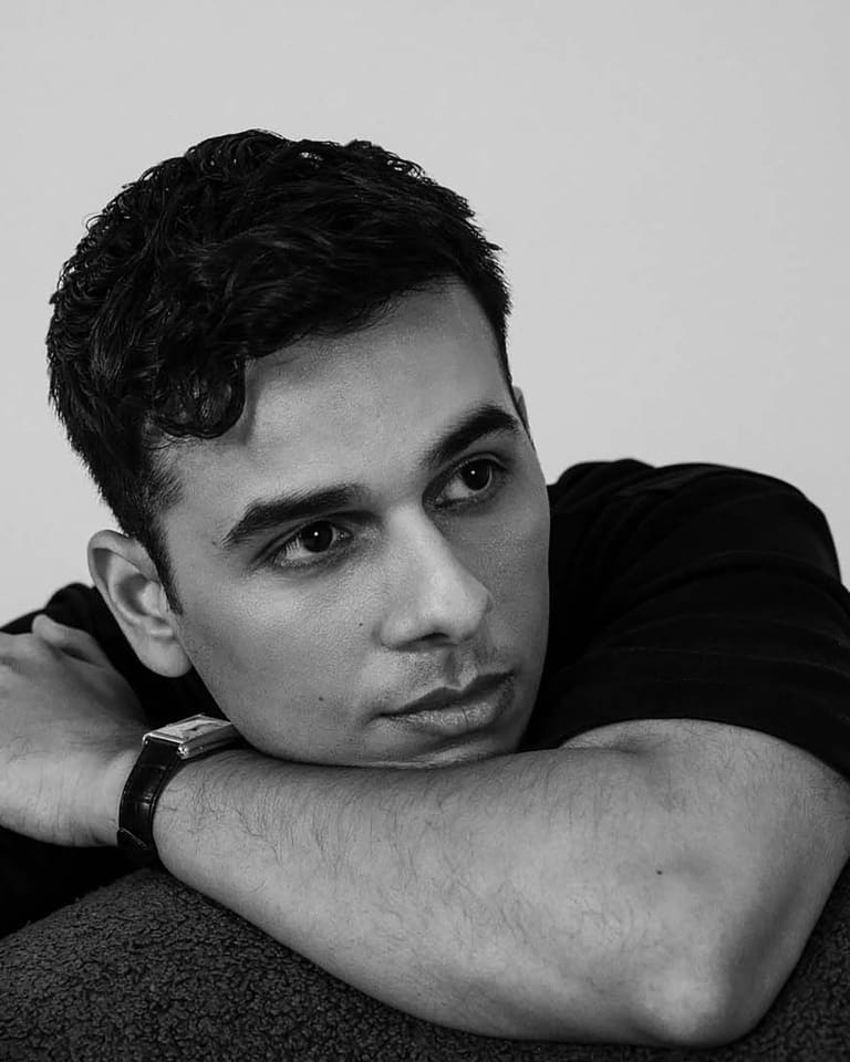Man with dark hair resting his chin on his arms against a neutral grey background in black and white portrait.