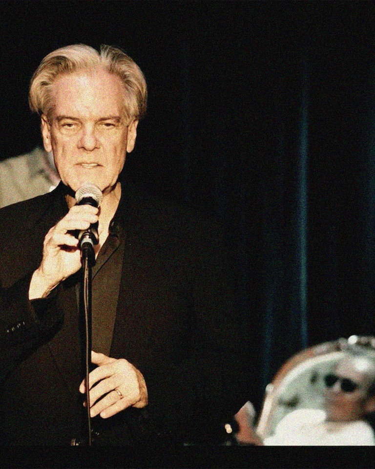 Older male singer in dark suit holding microphone centre stage, with band members visible either side.