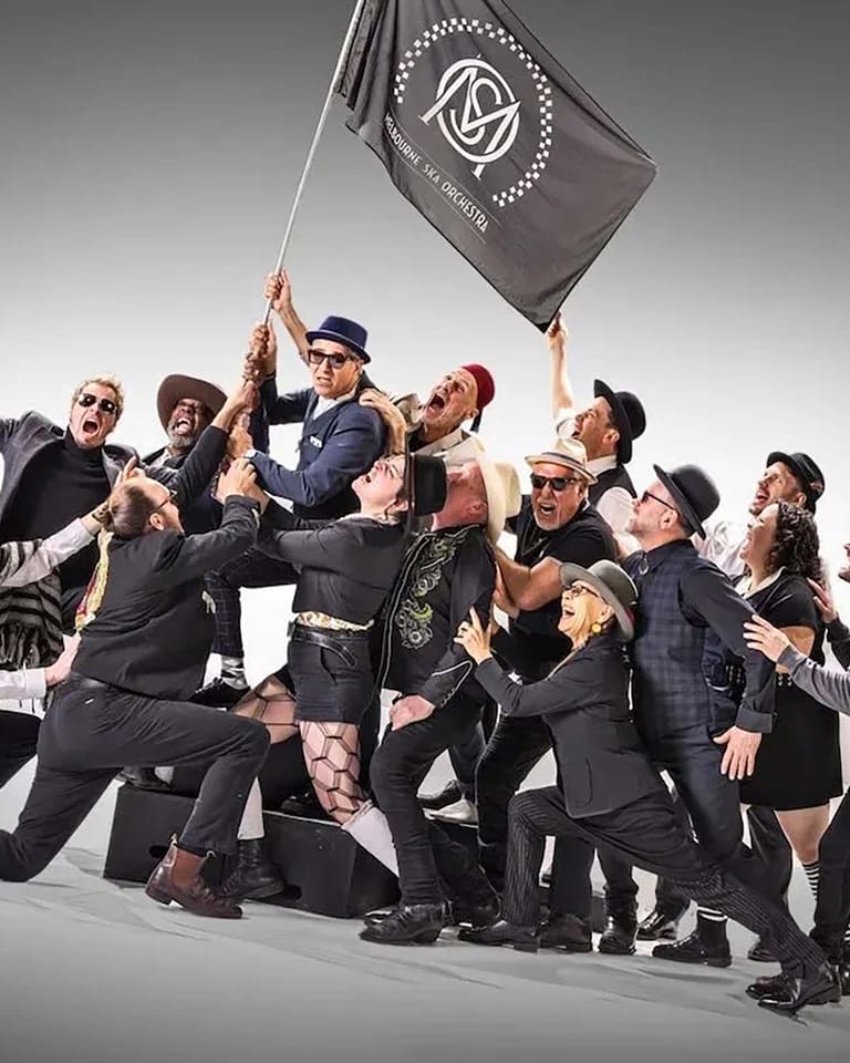 Group of performers in dark suits clustered together, one holding aloft a grey flag, against a neutral background.