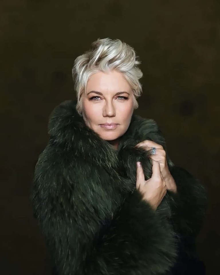 Person with platinum blonde hair wearing a black fur-collared coat against a dark background.