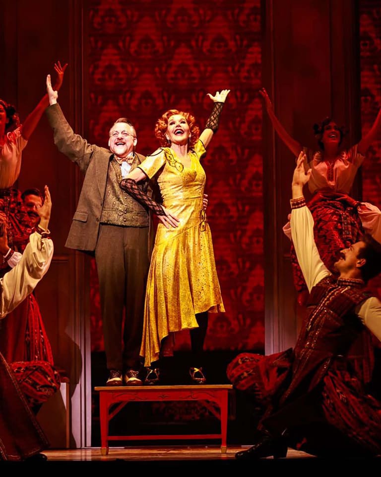 Two performers in vintage clothing stand centre stage with arms raised, surrounded by ensemble cast on a richly patterned red set.