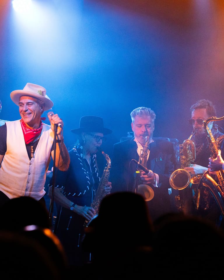 Band members in colourful stage outfits perform under blue and orange spotlights on a dark stage.